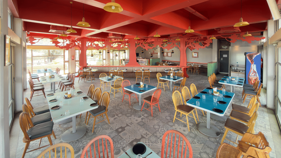The restaurant at Vinti Resort, Ambaji, warmly lit for dinner, features a striking red ceiling and comfortable tables set for dining.