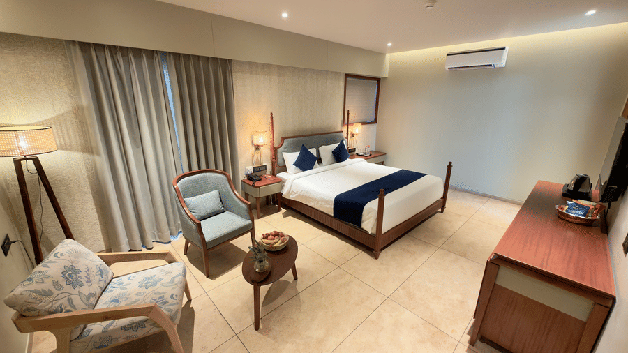 A well-lit, contemporary guest room at Vinti Resort, Ambaji shows a neat bed with plush pillows and a wall-mounted television.