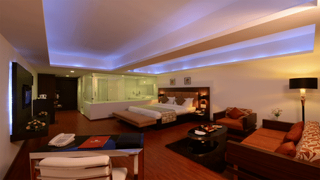 a king size bed room with wall-mount TV, a lounge area, a workstation, a minibar, and an attached see through washroom with glass doors and windows at Top3 Lords Resort Bhavnagar