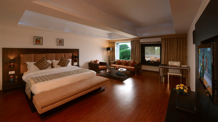 duplicate of a queen bedroom with air-conditioning, wall-mounted TV, workstation, a seating area with a sofa, and two separate glass windows showcasing the exterior lush greenery at Top3 Lords Resort Bhavnagar