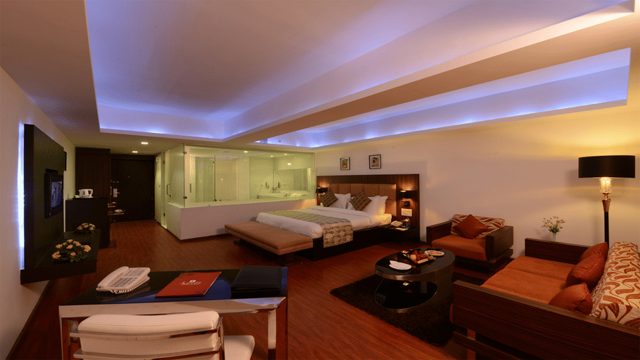 a king size bed room with wall-mount TV, a lounge area, a workstation, a minibar, and an attached see through washroom with glass doors and windows at Top3 Lords Resort Bhavnagar