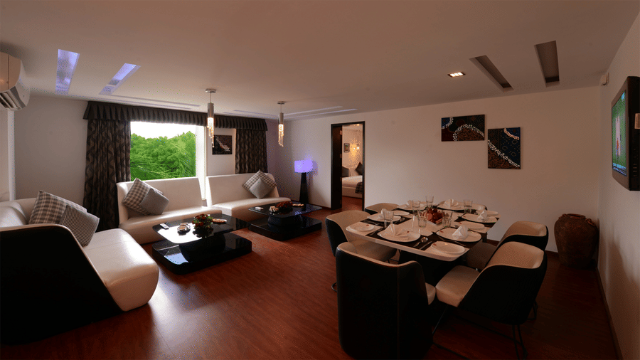 a separate lounge area of an executive king bed room featuring a seating area with sofas, an air-conditioner, a five seater dining area , and glass windows opening towards the lush greenery at Top3 LOrds Resort Bhavnagar