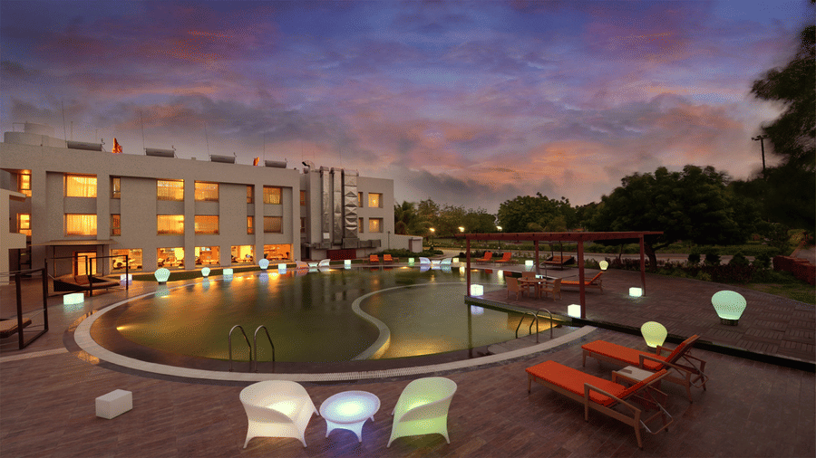 a large swimming pool surrounded by the Top3 Lords Resort Bhavnagar hotel's building, trees, ad sunloungers under a beautiful sky