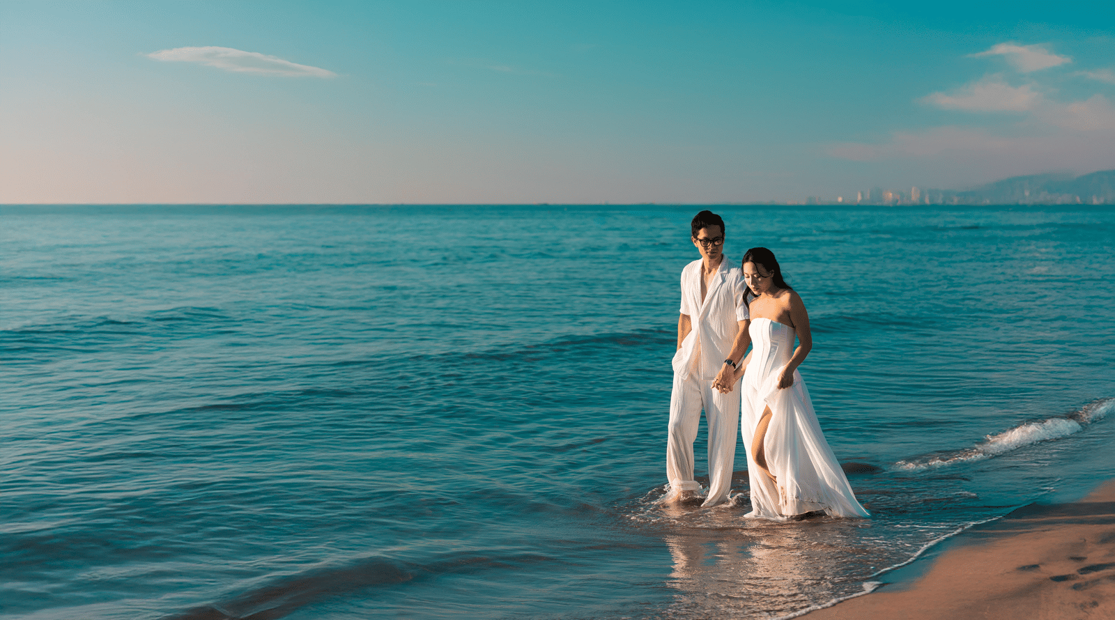 A couple in a white gown and suit standing in the gentle waves along the beach.