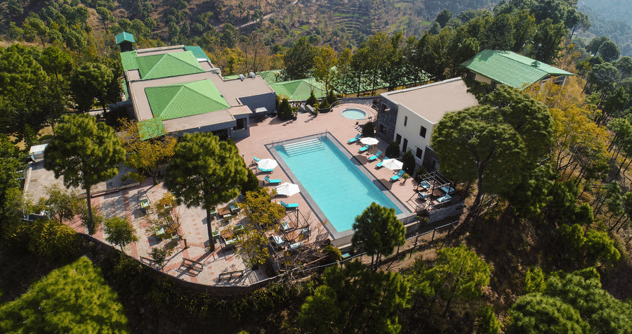 a large bird-eye shot of Suryavilas Luxury Resort and Spa featuring its swimming pool and premises surrounded by lush greenery on a hilltop