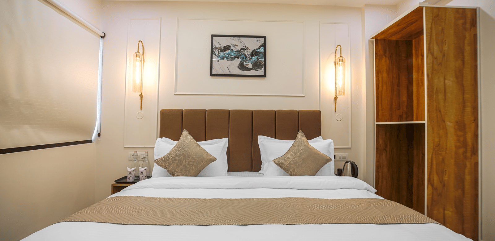 A neatly made king size bed at Perfectstayz Premium Varanasi, featuring a cushioned headboard, an artistic wall frame, and a wooden carved cupboard beside the bed.