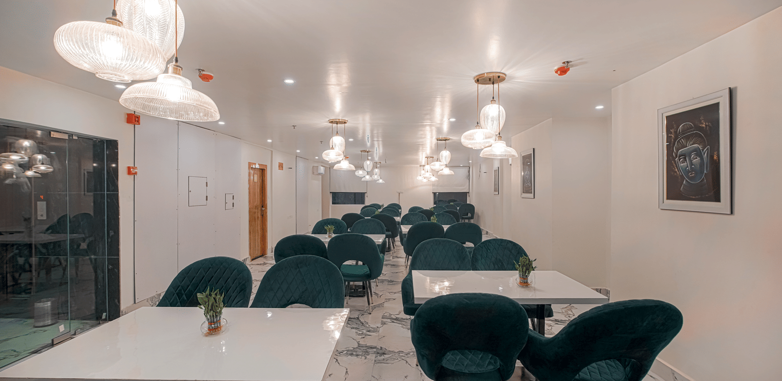 A clean, well-lit view of the modern dining room at Perfectstayz Premium Varanasi, featuring dark seating, white tables, an artistic wall frame, and marble flooring.