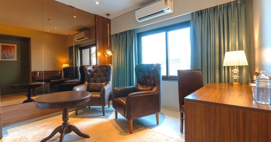 A seating area featuring a round wooden table, 2 leather armchairs, a desk with a lamp, and teal curtains by a window at Magnus Kapitol, Sakore Nagar, Viman Nagar.