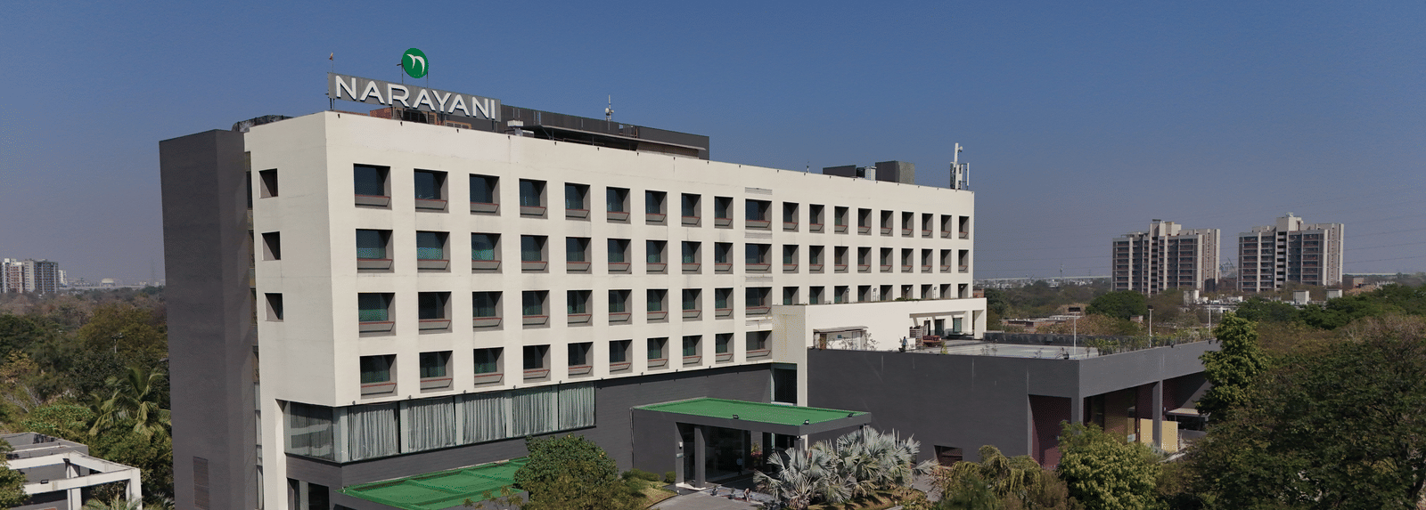 Long-shot of Narayani Heights Hotel & Resorts, Ahmedabad facade with trees surrounding it under a clear sky.