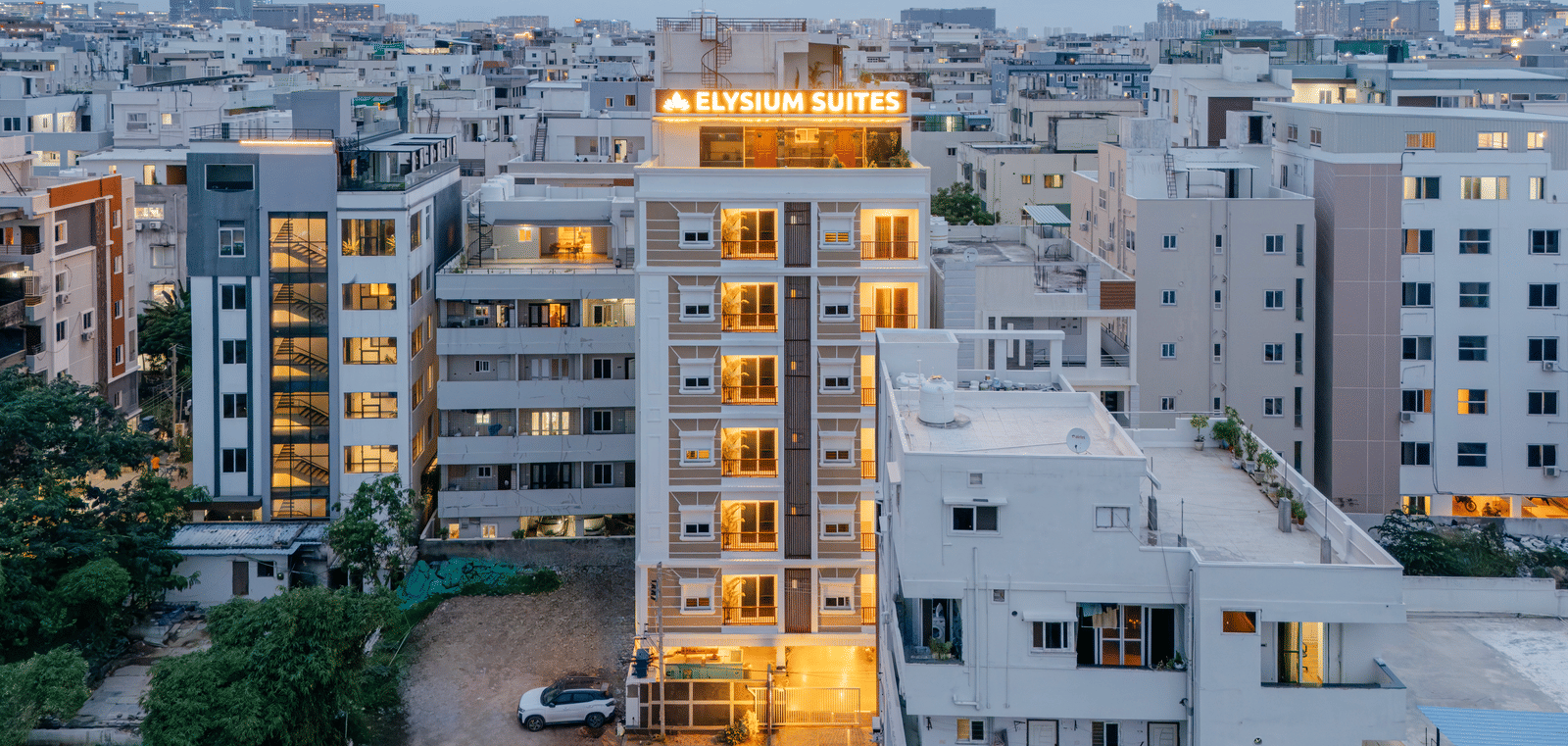 Buildings stand close together in a city at dusk with lights visible from windows as night falls near Elysium Studio Suites, Madhapur..