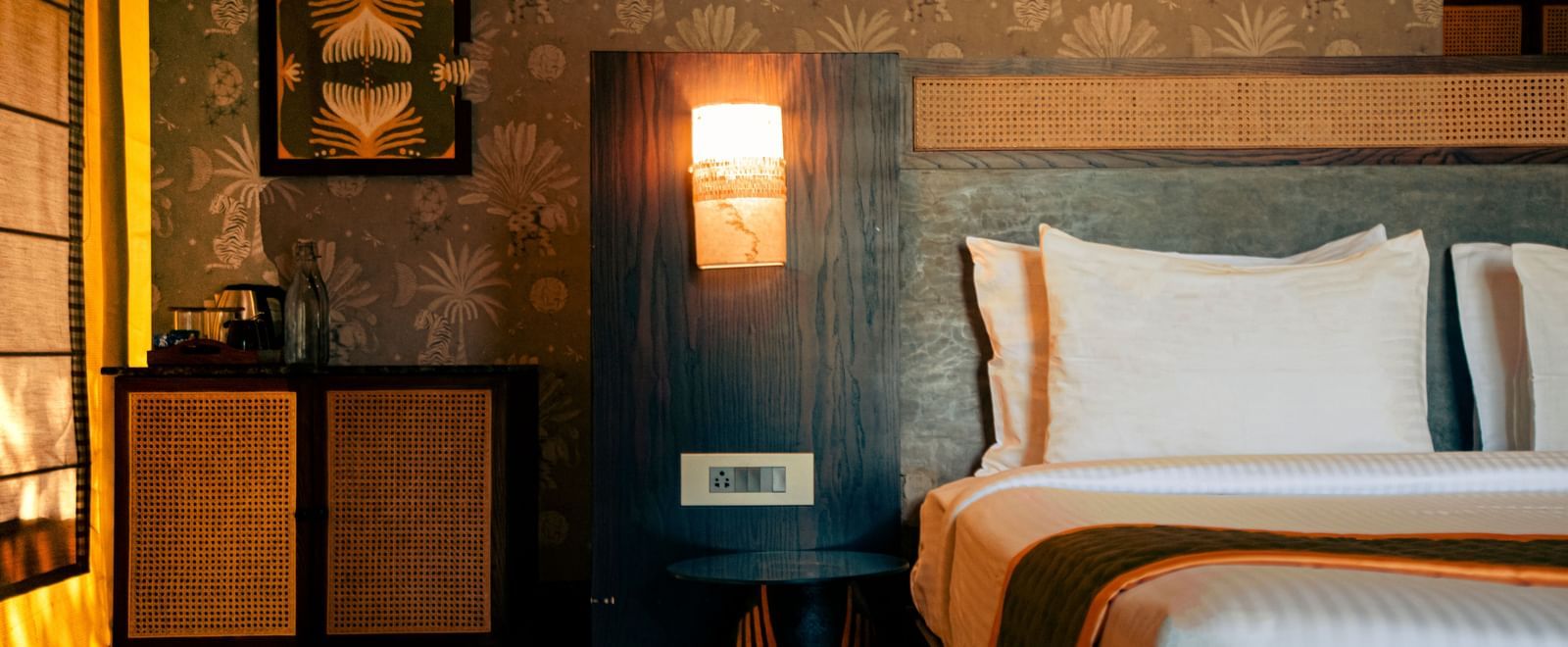Partially visible bed with wall painting, bedside lights and a small cupboard at Fireflies Resorts, Kabini.
