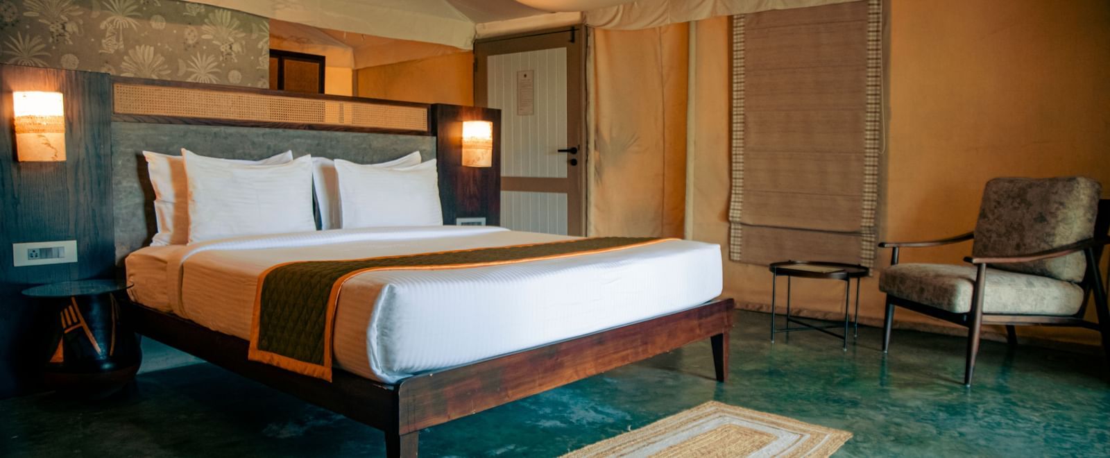 Room with king size bed, two lamps, chair, desk, and sloped ceiling at Fireflies Resorts, Kabini.