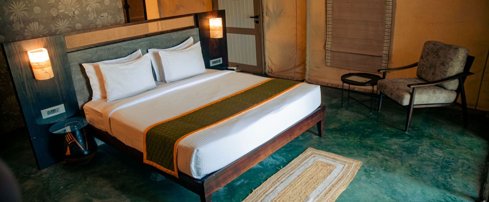 Room with king size bed, two bedside lamps, chair, and green floor at Fireflies Resorts, Kabini.