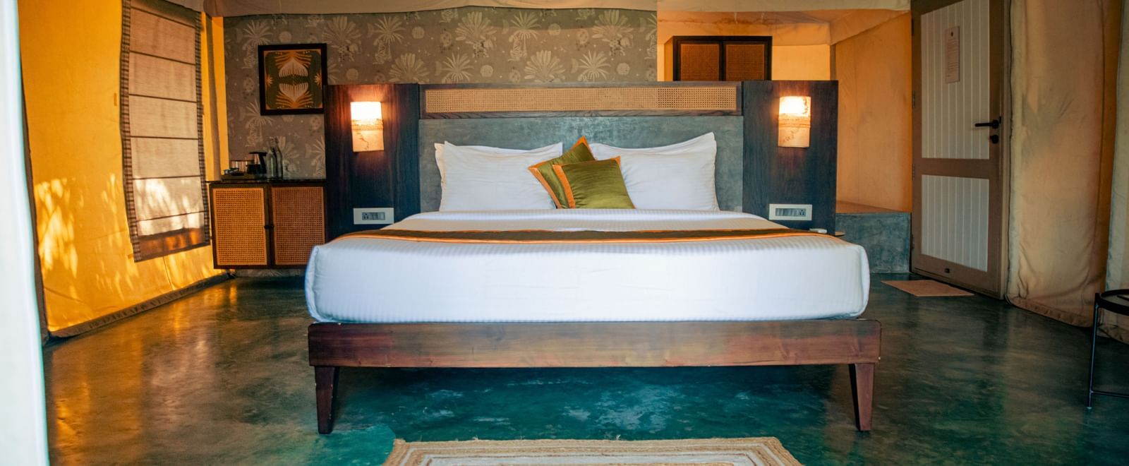 Bedroom with king size bed, green floor, two yellow cushions, nightstands with lamps, and a rug at Fireflies Resorts, Kabini.