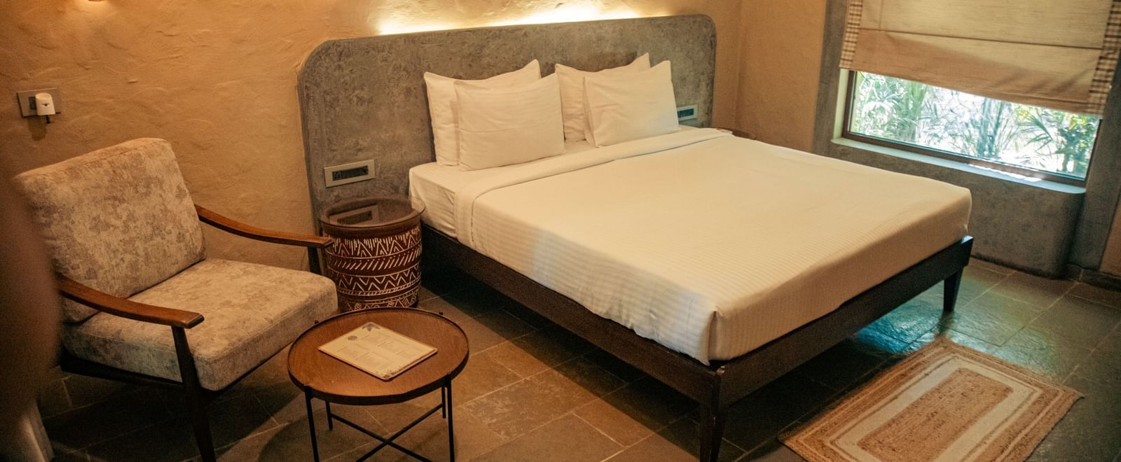 Room with a king size bed, two chairs, a round side table, and beige walls at Fireflies Resorts, Kabini.