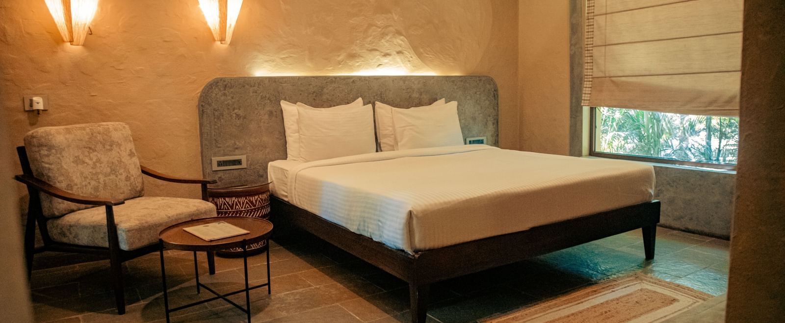 Cosy bedroom with a king size bed, an armchair, side table and wall lights against a soft beige wall at Fireflies Resorts, Kabini.