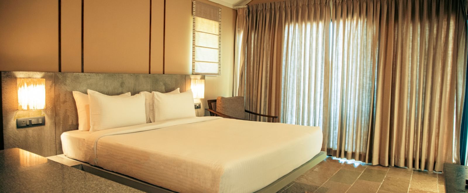 Bedroom with king size bed, bedside lamps, cushioned headboard, and full-length curtain covering a large window at Fireflies Resorts, Kabini.