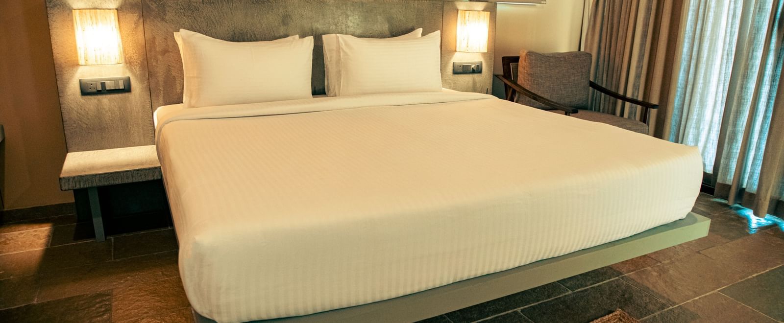 Bedroom with king size bed, bedside lamps, floor rug, and sheer curtains covering a glass window at Fireflies Resorts, Kabini.