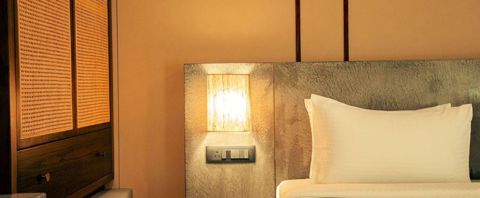 Close-up of a neatly made bed with a cushioned headboard, bedside light, and panelled wall at Fireflies Resorts, Kabini.
