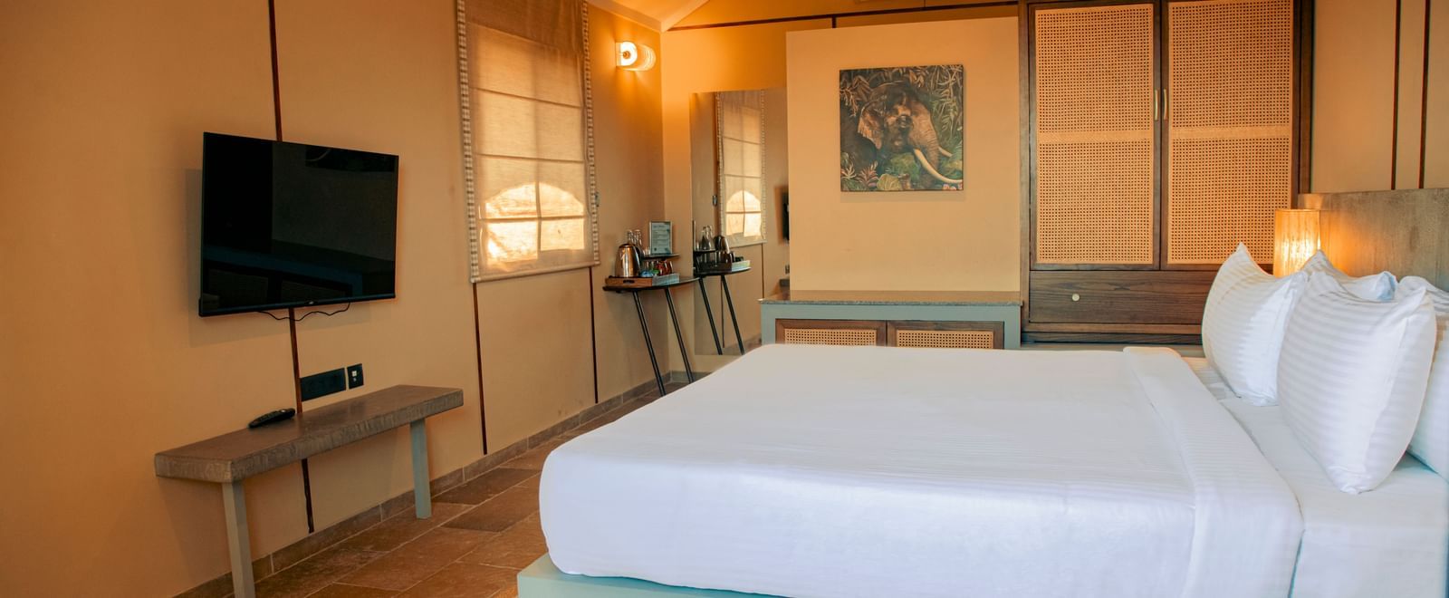 Room with a king size bed, mounted TV,  bedside lamps, and beige panelled walls at Fireflies Resorts, Kabini.