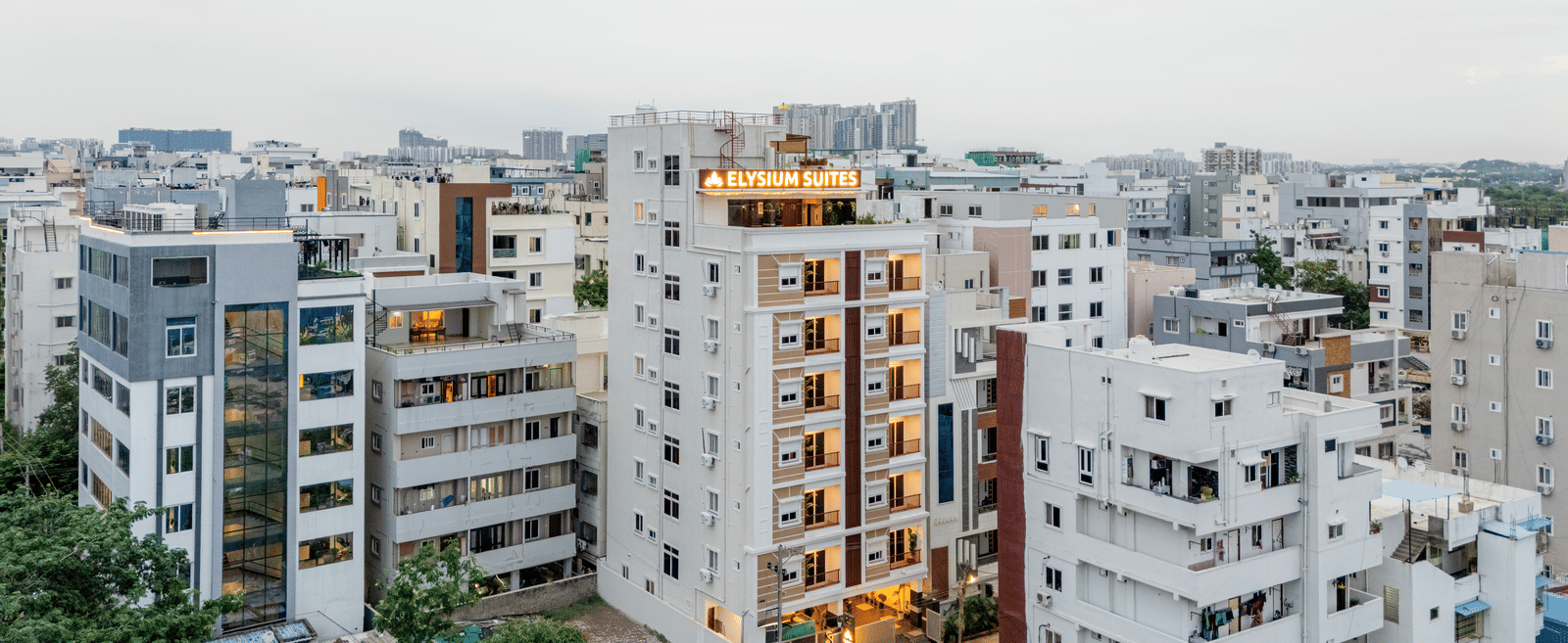 Buildings stand close together in a city under a bright sky with lights visible from windows near Elysium Studio Suites, Madhapur.