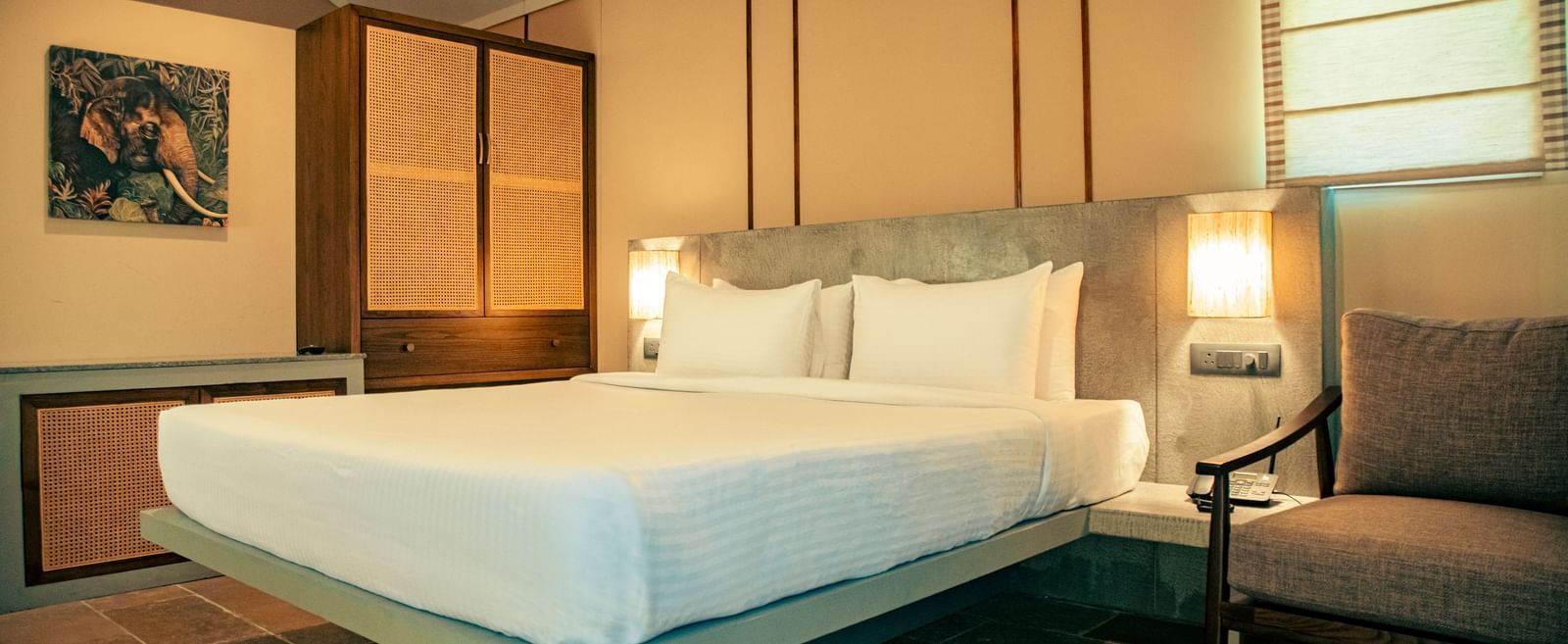 Room with wooden-panelled walls, king size bed with white bedding, two lamps, chair, and wall-mounted painting at Fireflies Resorts, Kabini.