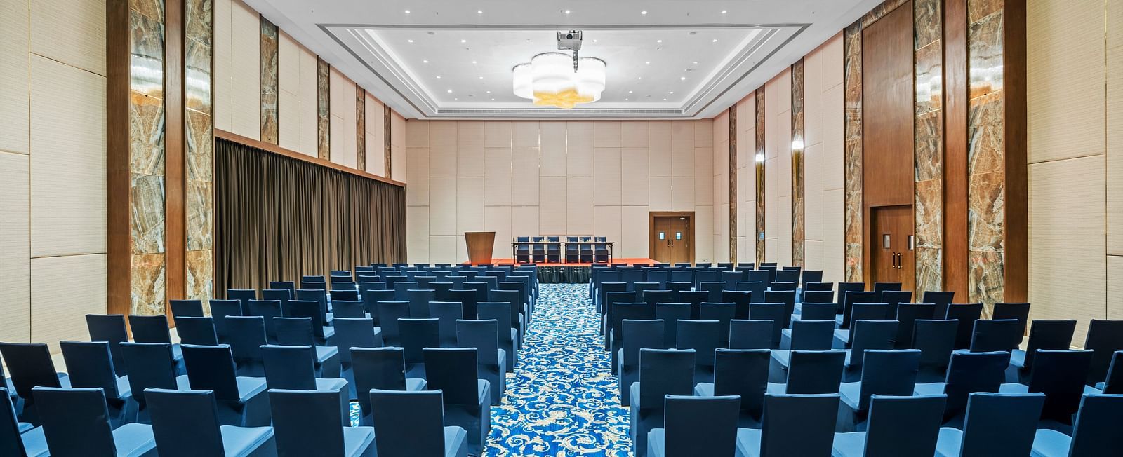 An image of Champavati Banquet Hall with blue chairs facing towards a podium at Grand Vijayawada by GRT Hotels.