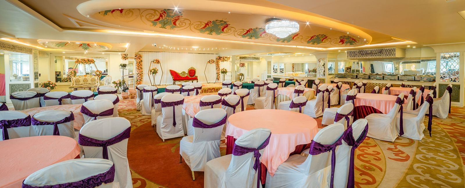 Samrat featuring round tables draped in pink tablecloths and chairs adorned with white covers and purple sashes.