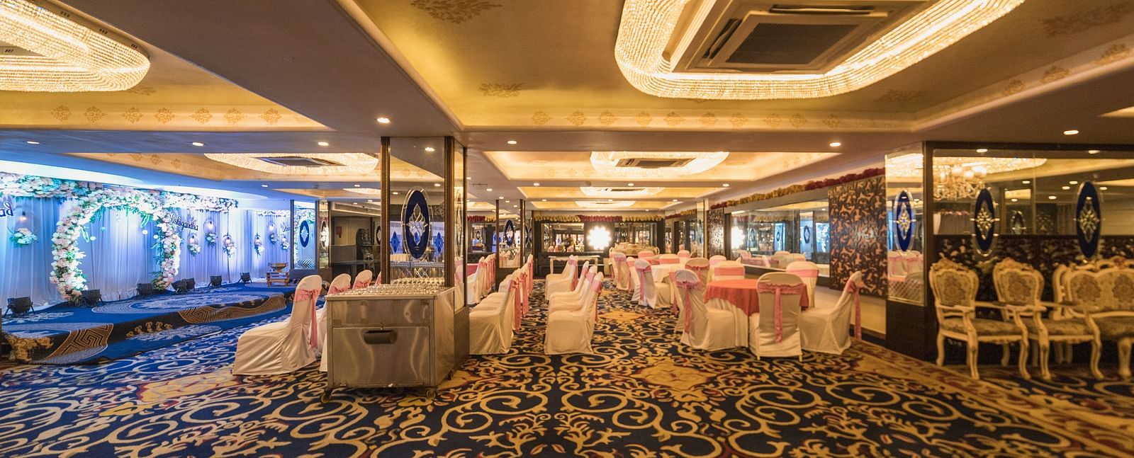Magadh with a patterned blue carpet, tables set with white linens, and modern lighting fixtures.