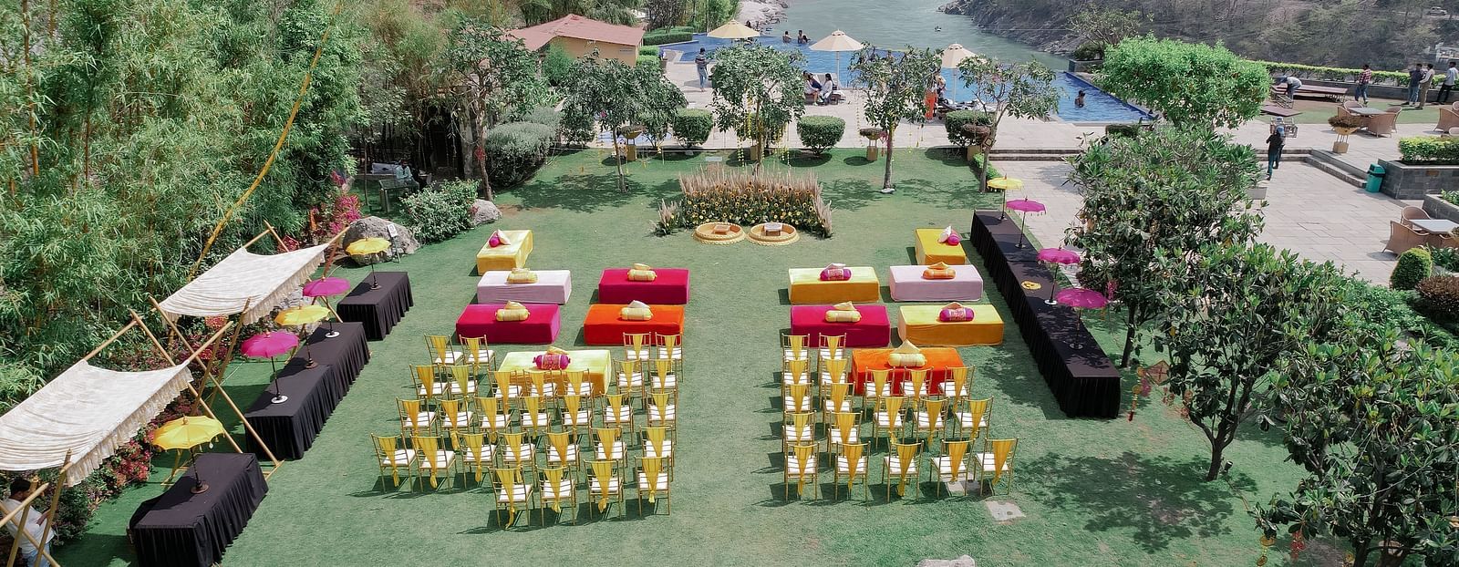 Aerial view of the outdoor venue. The lawn is decorated with colorful seating arrangements, including rows of chairs and square seating platforms. A large floral decor setup is visible in the center