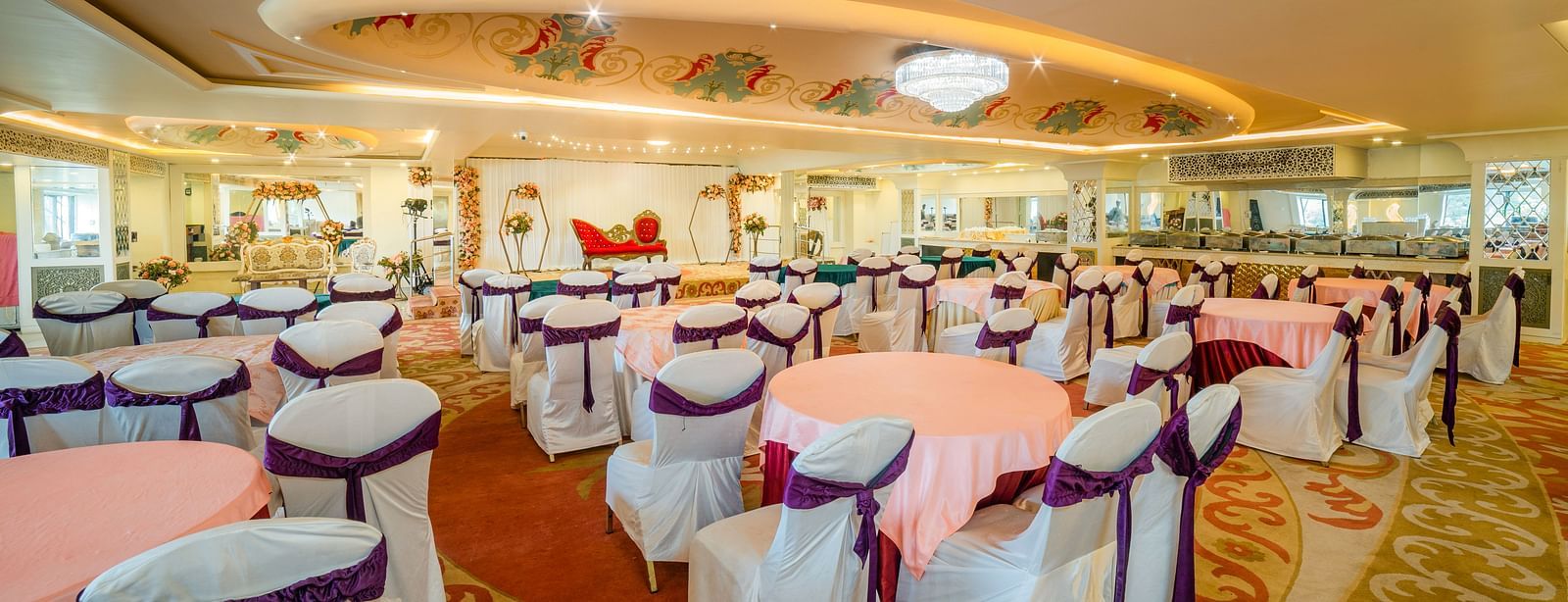Samrat featuring round tables draped in pink tablecloths and chairs adorned with white covers and purple sashes.