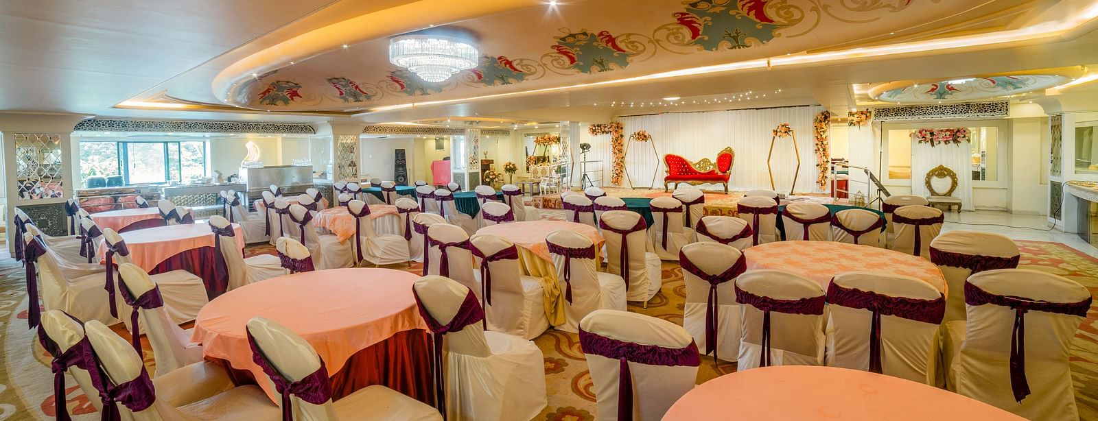 Samrat featuring round tables with pastel-colored tablecloths and cushioned chairs, under warm lighting and decorative ceiling elements.