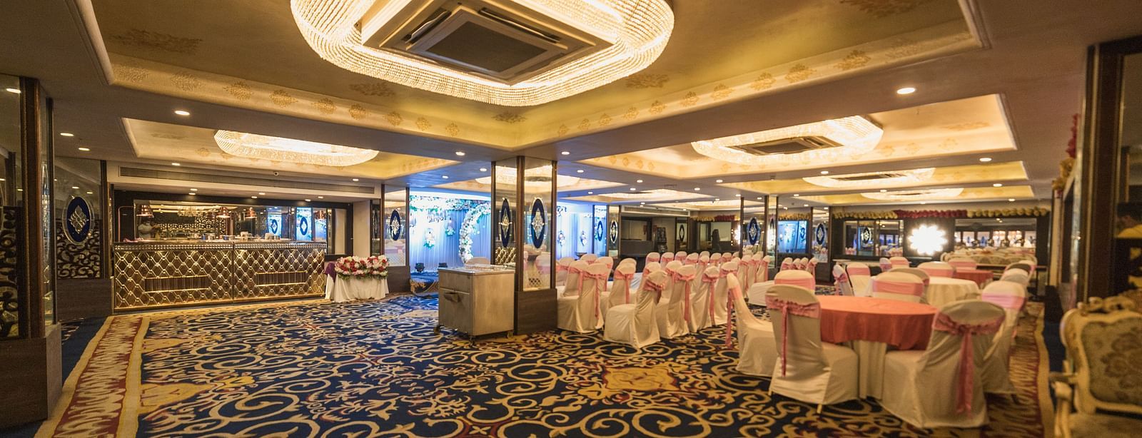 Magadh with round tables, chairs with covers, patterned carpeting, and decorative lighting fixtures on the ceiling.
