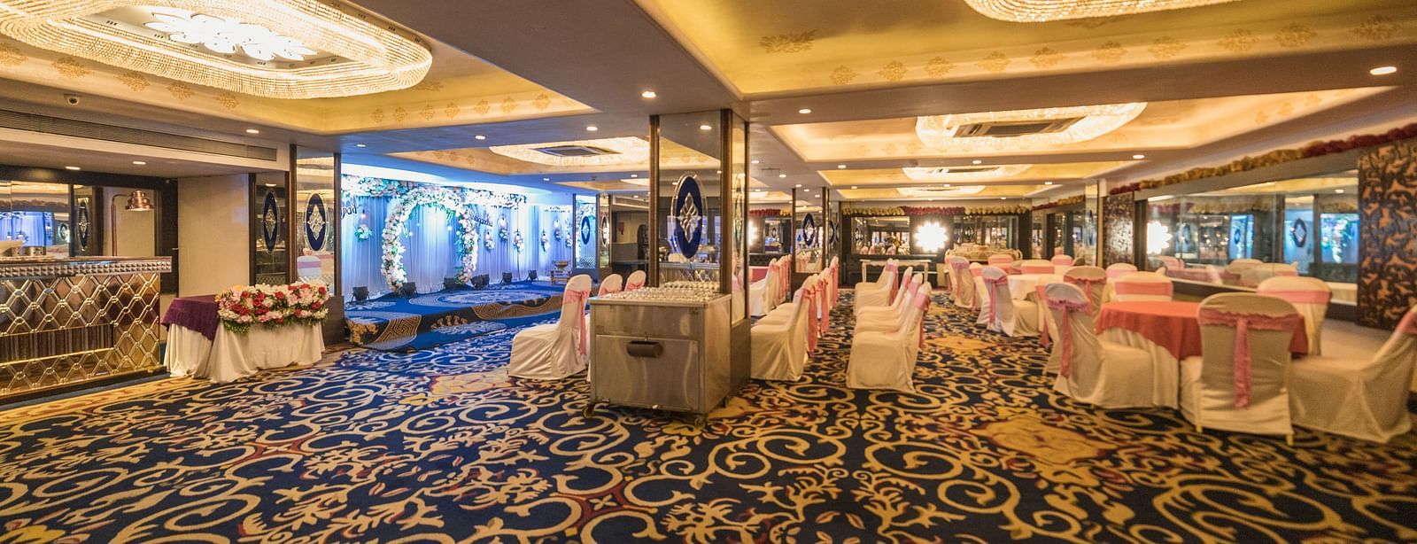 Magadh featuring shows tables draped in white cloths, chairs with pink accents, and a patterned blue carpet.