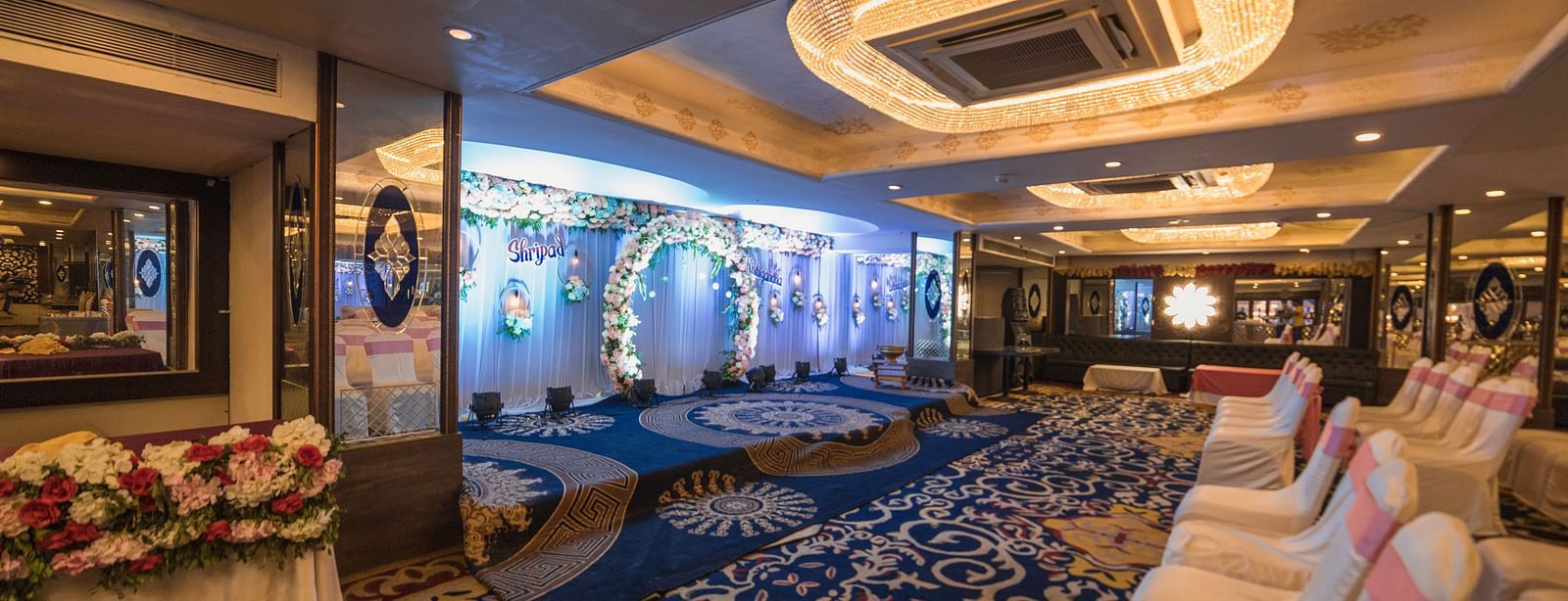 Magadh showcasing blue patterned carpeting, chairs with pink and white covers, and modern ceiling lights.