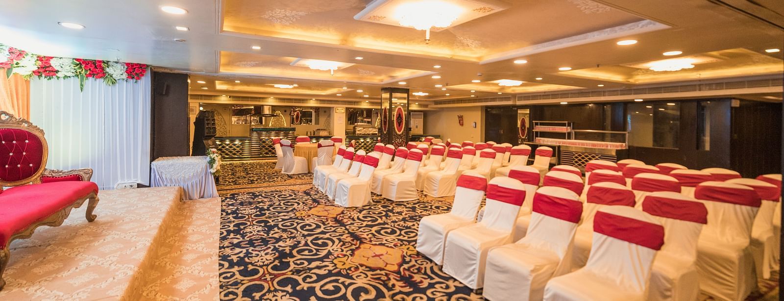 Maurya featuring rows of chairs covered in white fabric and red sashes, and a patterned carpet.