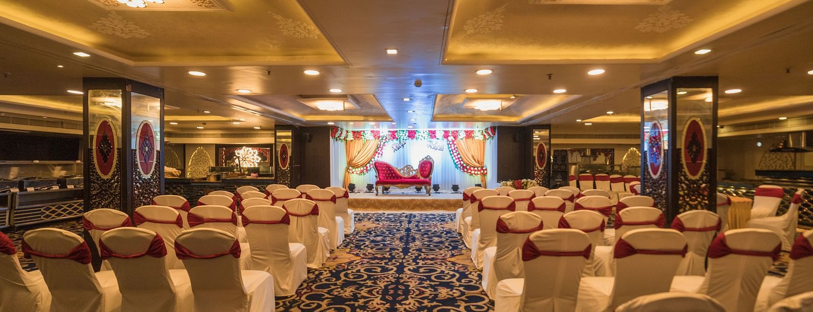 Maurya showcasing a arrangement of white-covered chairs with red sashes, a patterned carpet, and a stage area with comparable decor and lighting.