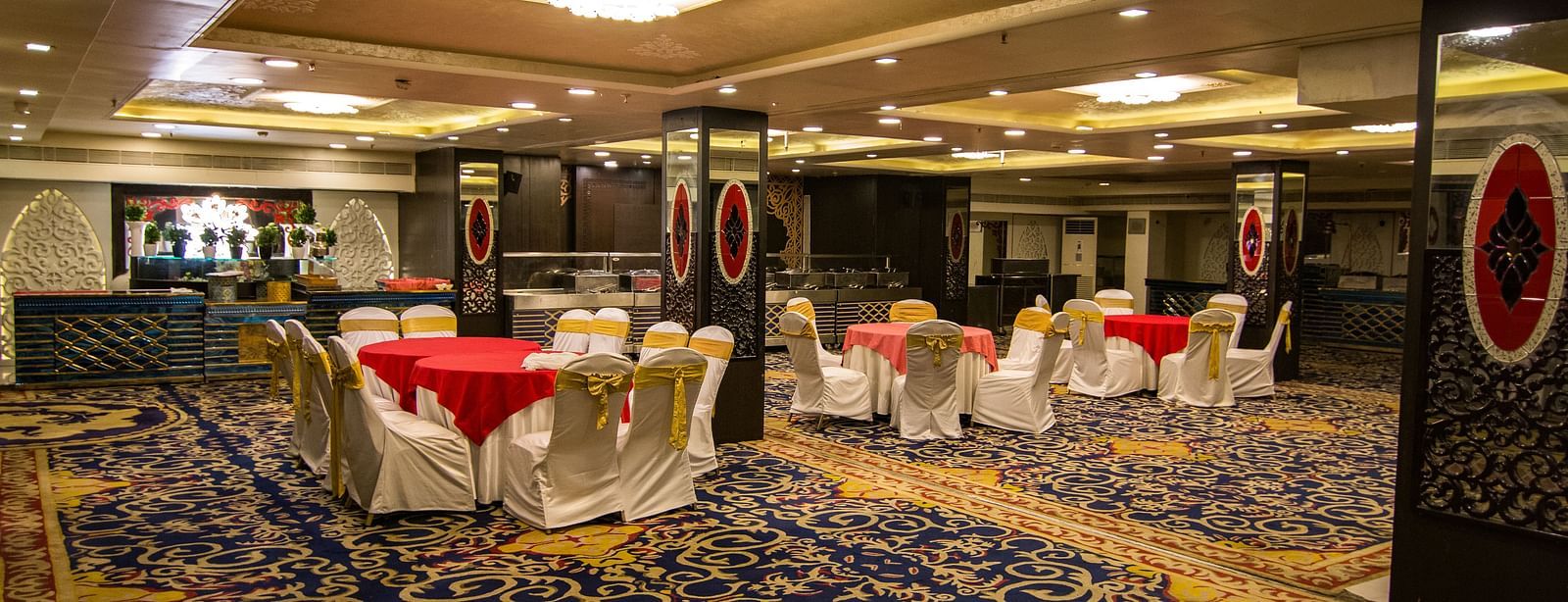 Maurya featuring round tables draped in red cloths and surrounded by white chairs, featuring a patterned carpet and decorative pillars.