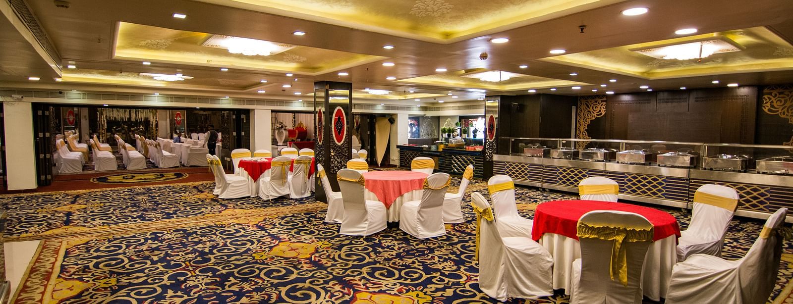 Maurya featuring round tables covered in red cloths and surrounded by white chairs, under a well-lit ceiling and patterned carpet.