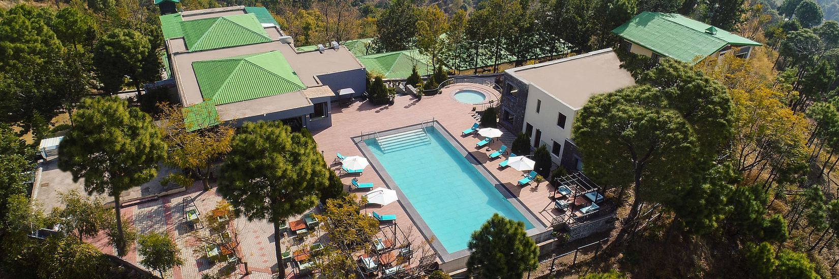 a large bird-eye shot of Suryavilas Luxury Resort and Spa featuring its swimming pool and premises surrounded by lush greenery on a hilltop