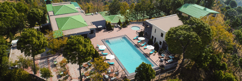 a large bird-eye shot of Suryavilas Luxury Resort and Spa featuring its swimming pool and premises surrounded by lush greenery on a hilltop