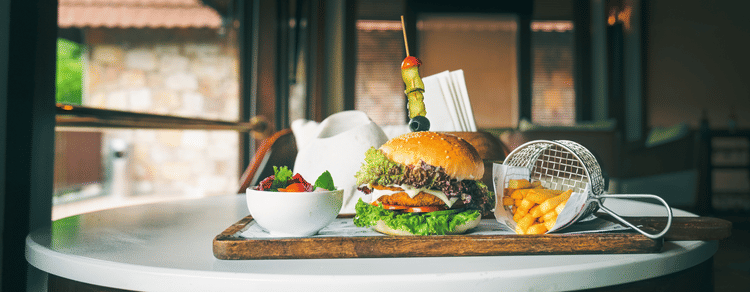 A gourmet burger served with a side of fries in a mini fryer basket and a fresh salad, presented on a wooden board in a cosy indoor restaurant setting - Ananta Spa and Resort, Pushkar.