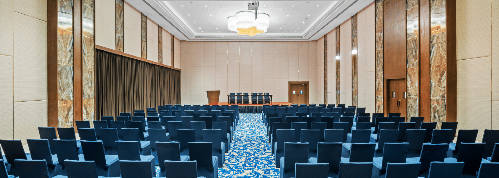 An image of Champavati Banquet Hall with blue chairs facing towards a podium at Grand Vijayawada by GRT Hotels.