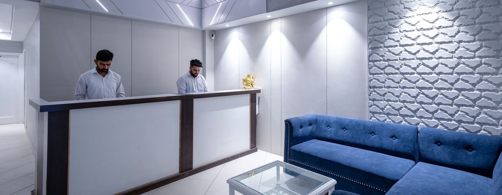 The reception area with two staff members dressed in white formal shirts standing behind the reception desk - Hotel Downtown17, Chandigarh.