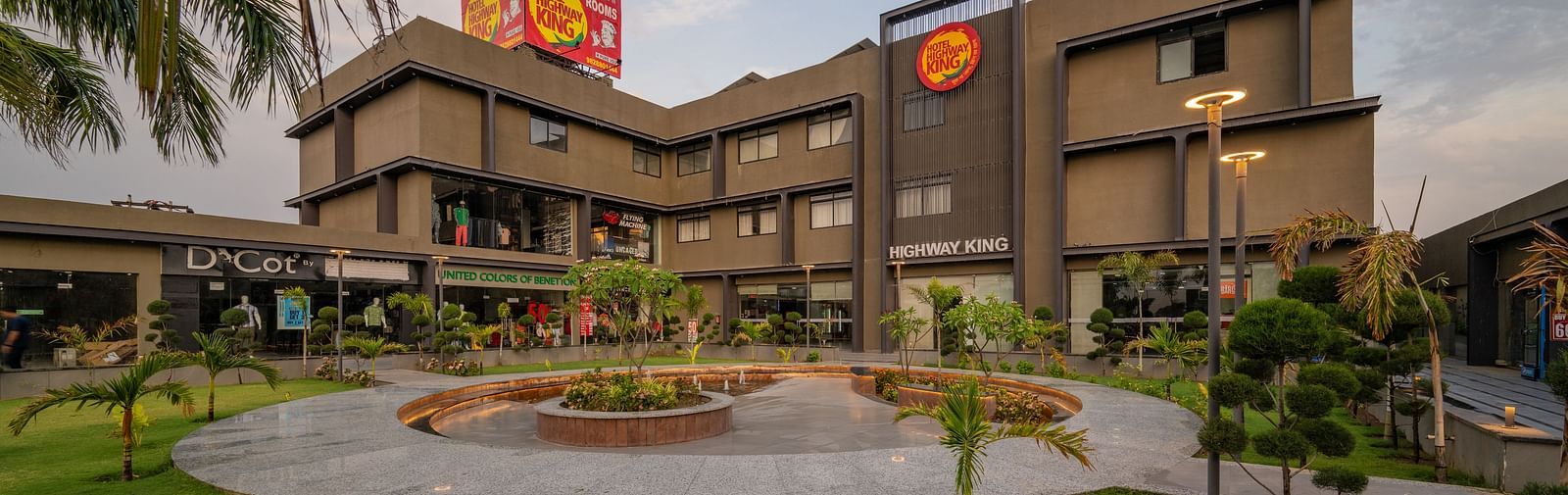A modern, multi-storey building with a distinctive facade is surrounded by a landscaped garden with a circular fountain in the foreground.