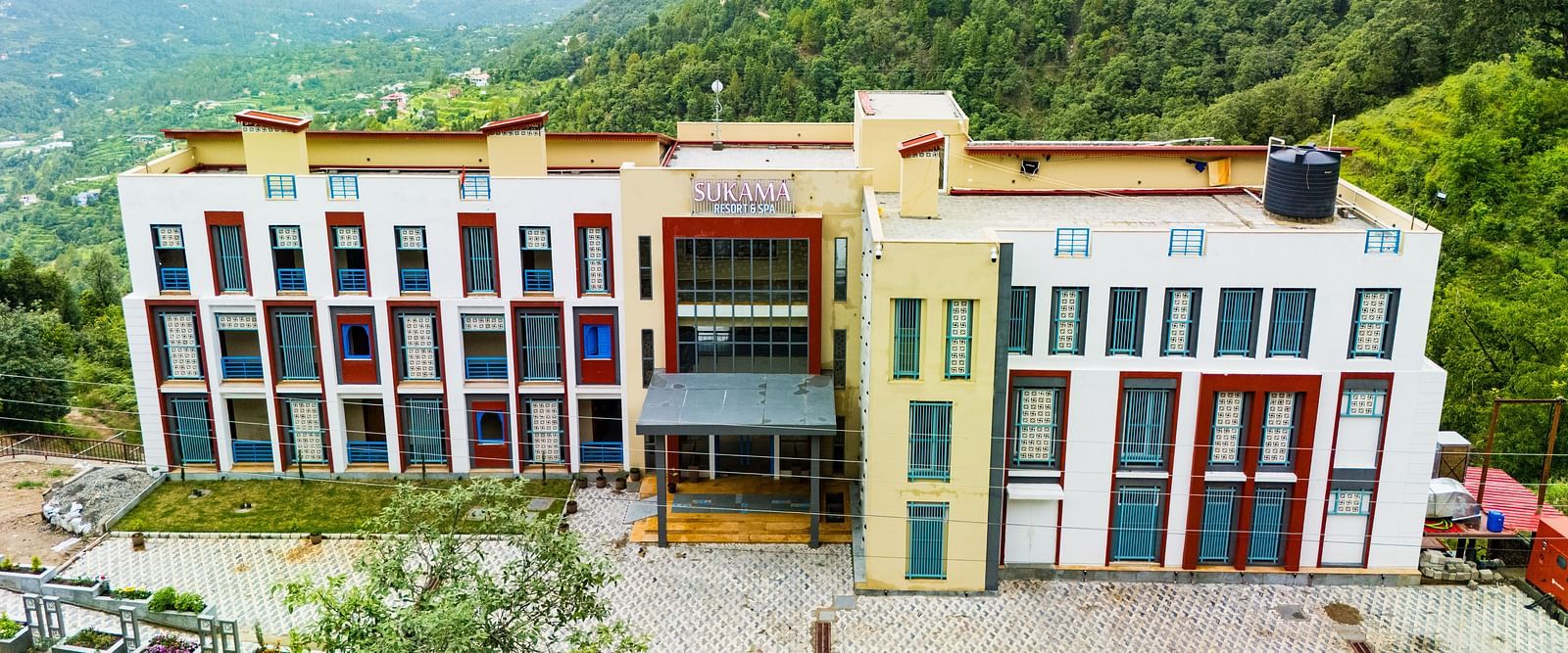 A colourful building complex is nestled in a mountainous, green landscape under a bright sky | Sukama Resort & Spa, Nainital