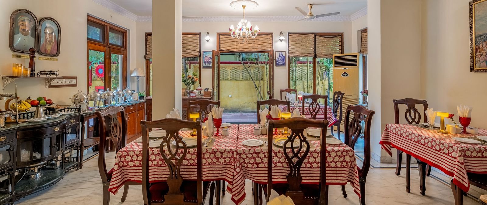 The spacious dining hall at Dera Rawatsar, Jaipur, offers set tables with patterned cloths, a buffet setup, and a view towards an outdoor area.
