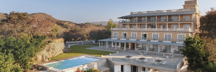 A facade of Fateh Safari Resort in Kumbhalgarh featuring a green lawn and a patio with tables and chairs. It also features a swimming pool set amidst the trees.