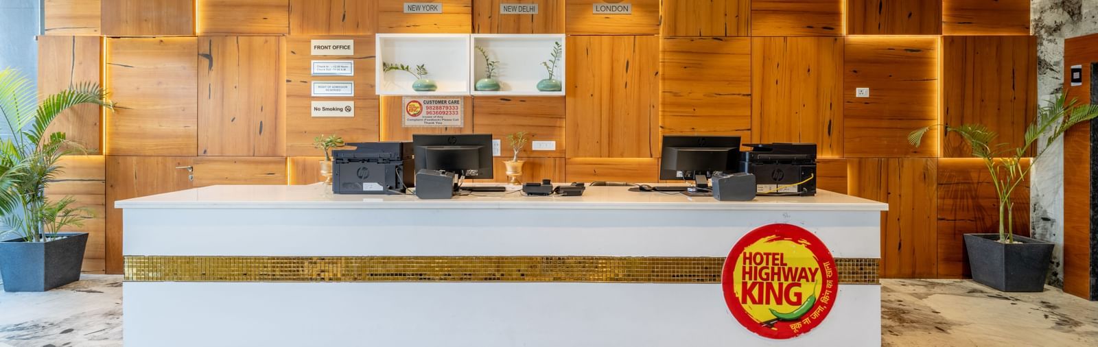 Reception desk at Hotel Highway King, Bagru, with computer stations, wooden paneled walls, and hanging light fixtures.