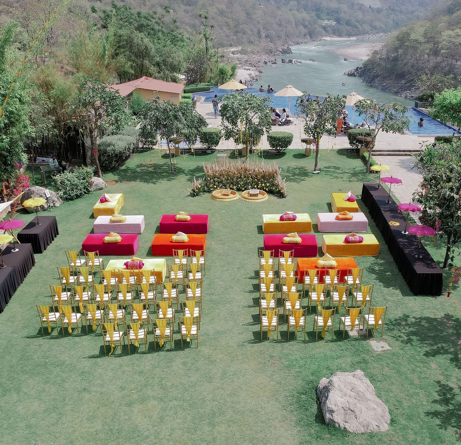 Aerial view of the outdoor venue. The lawn is decorated with colorful seating arrangements, including rows of chairs and square seating platforms. A large floral decor setup is visible in the center